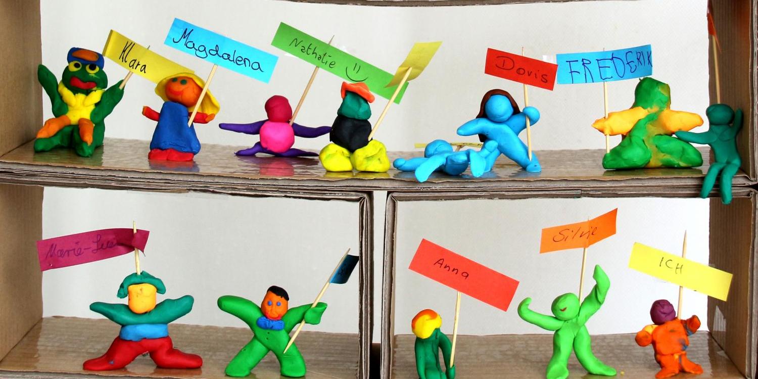Eine bunte Gruppe aus Knetfiguren hält kleine Namensschilder und Fähnchen in farbenfrohen Kartonfächern hoch.