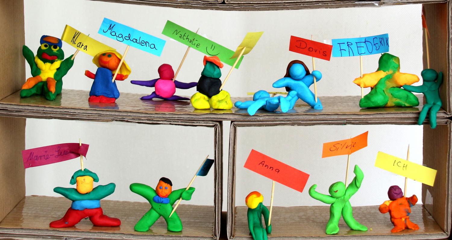 Eine bunte Gruppe aus Knetfiguren hält kleine Namensschilder und Fähnchen in farbenfrohen Kartonfächern hoch.