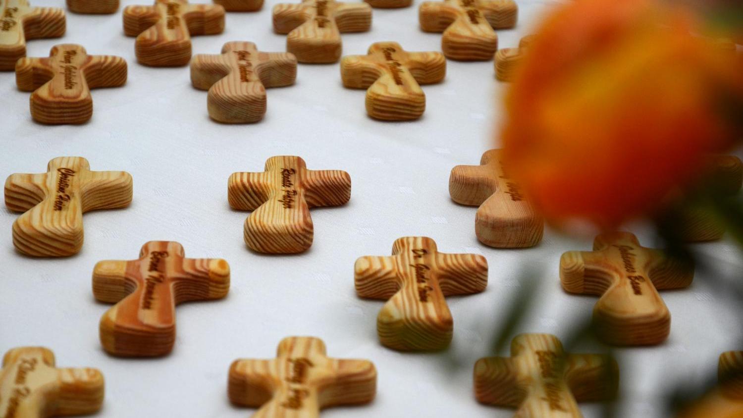 Mehrere kleine Holzkreuze liegen auf weißem Untergrund, rechts vorne eine orange Blume.
