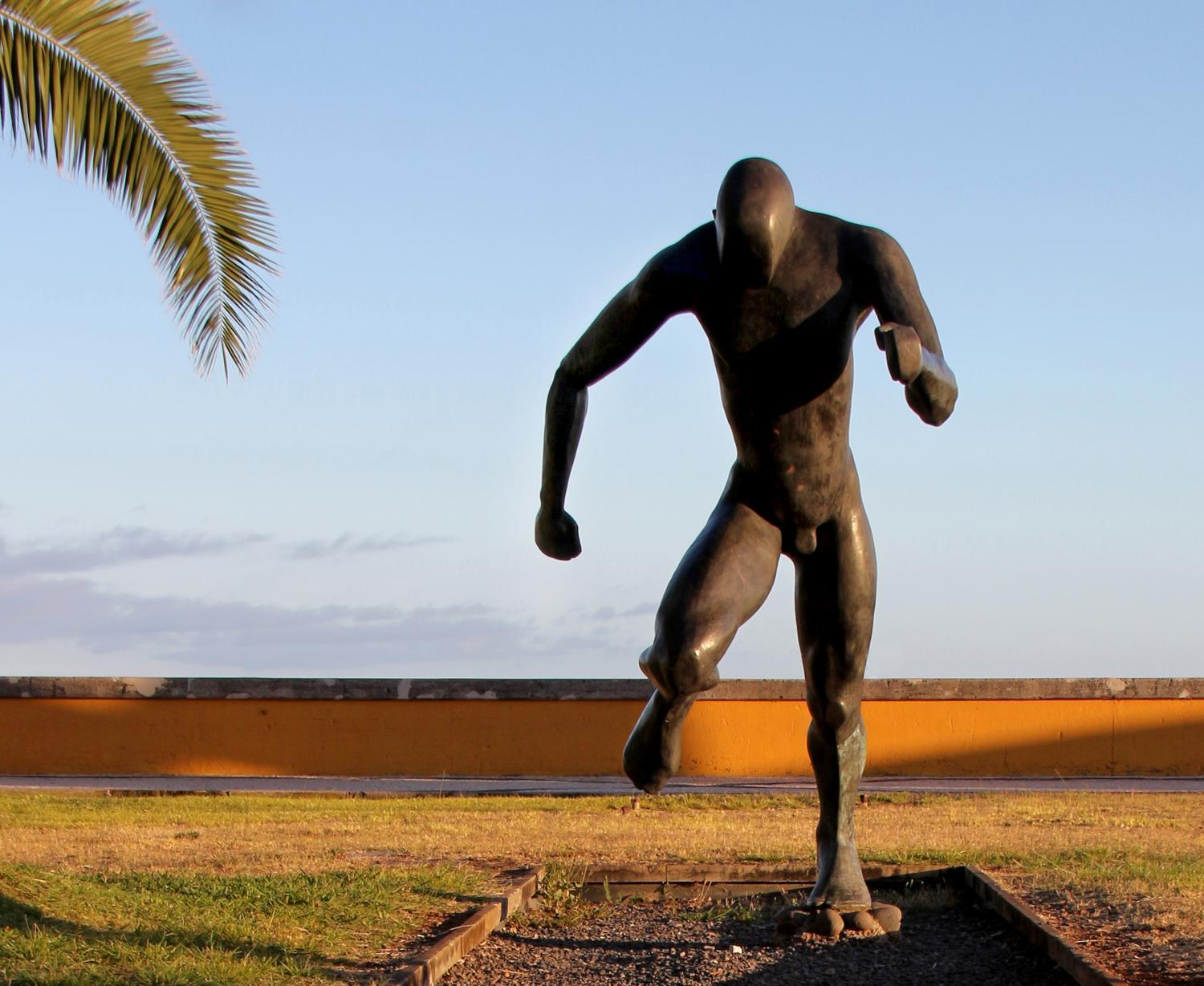 Eine dunkle Bronzeskulptur zeigt eine menschliche Figur in Laufhaltung auf einem kleinen Sockel im Freien, mit Gras und einer Palme im Hintergrund.