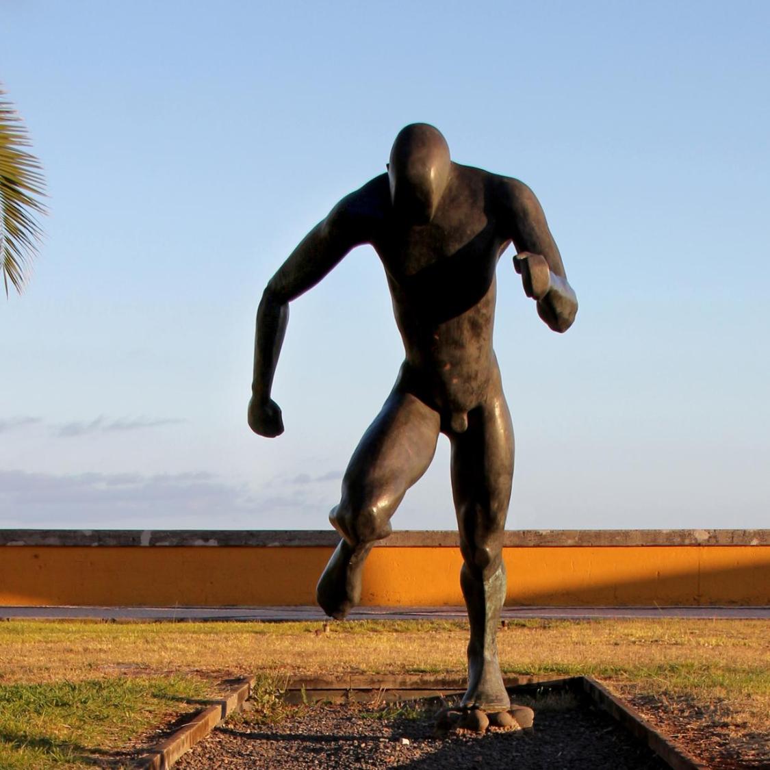 Eine dunkle Bronzeskulptur zeigt eine menschliche Figur in Laufhaltung auf einem kleinen Sockel im Freien, mit Gras und einer Palme im Hintergrund.