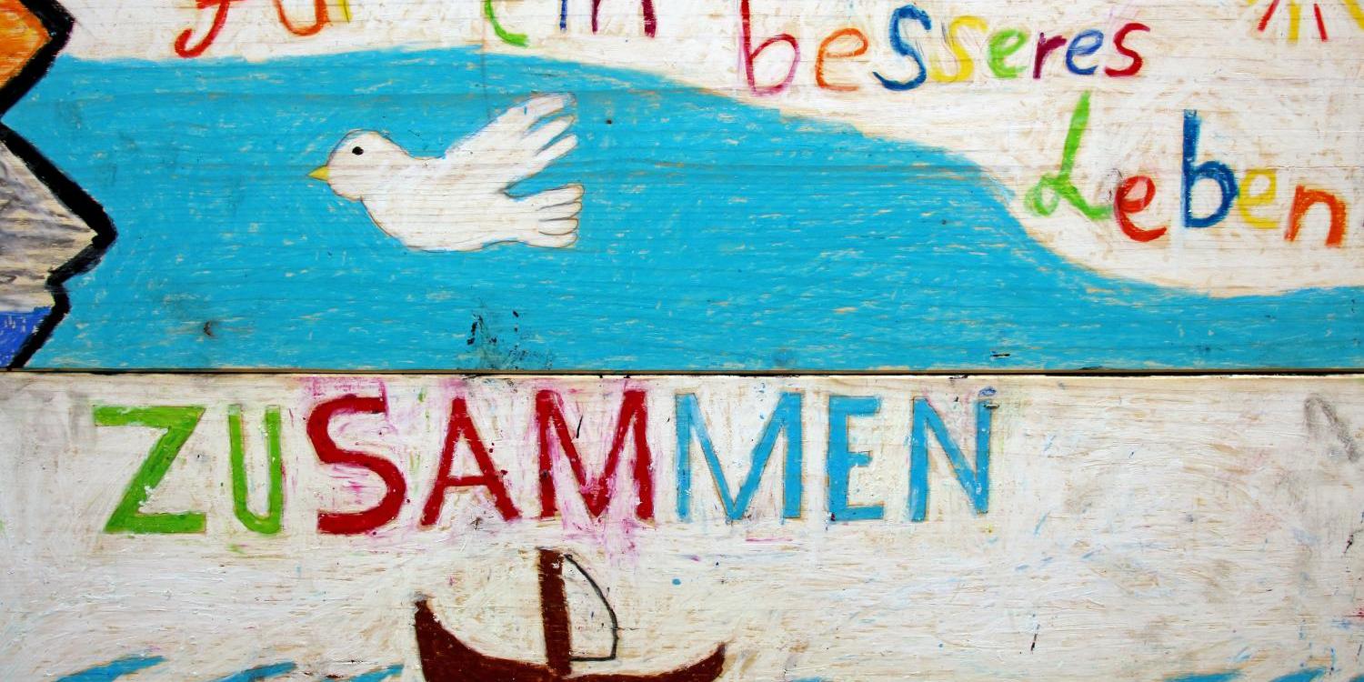 Ein bunt gemaltes Bild zeigt eine weiße Friedenstaube über blauem Wasser, darunter ein kleines Boot auf Wellen, begleitet vom Schriftzug „Für ein besseres Leben zusammen“.