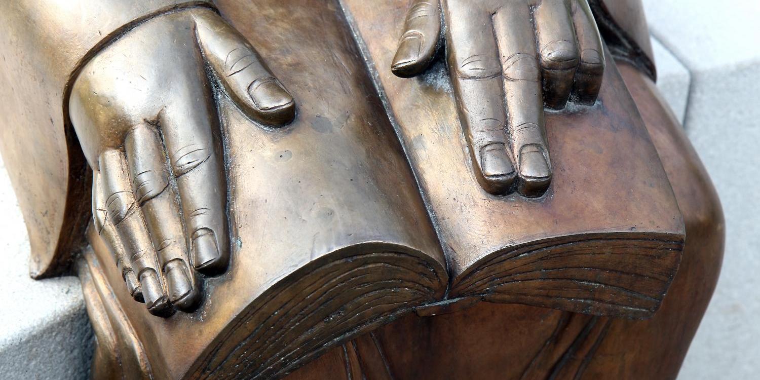 Eine bronzene Skulptur zeigt zwei Hände, die auf einem geöffneten Buch ruhen.