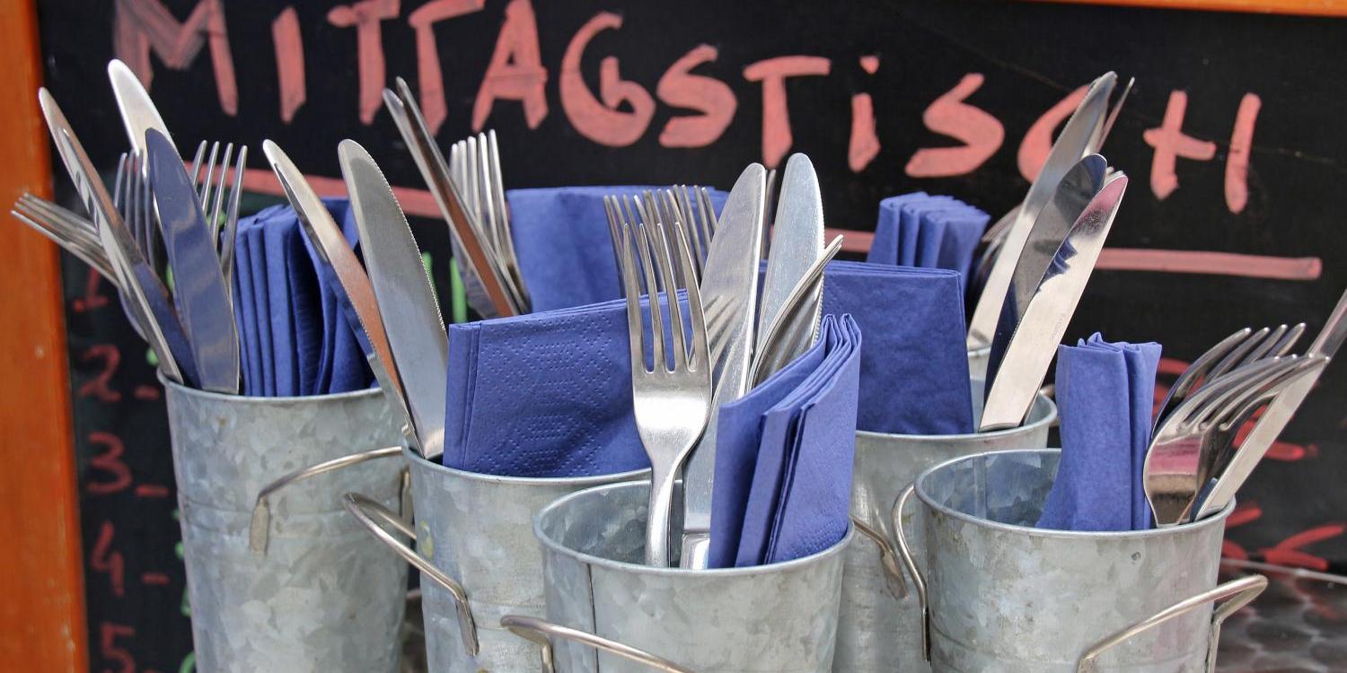 Mehrere Metallbecher sind mit Besteck und blauen Servietten gefüllt, davor steht eine Tafel mit der Aufschrift „Mittagstisch“.