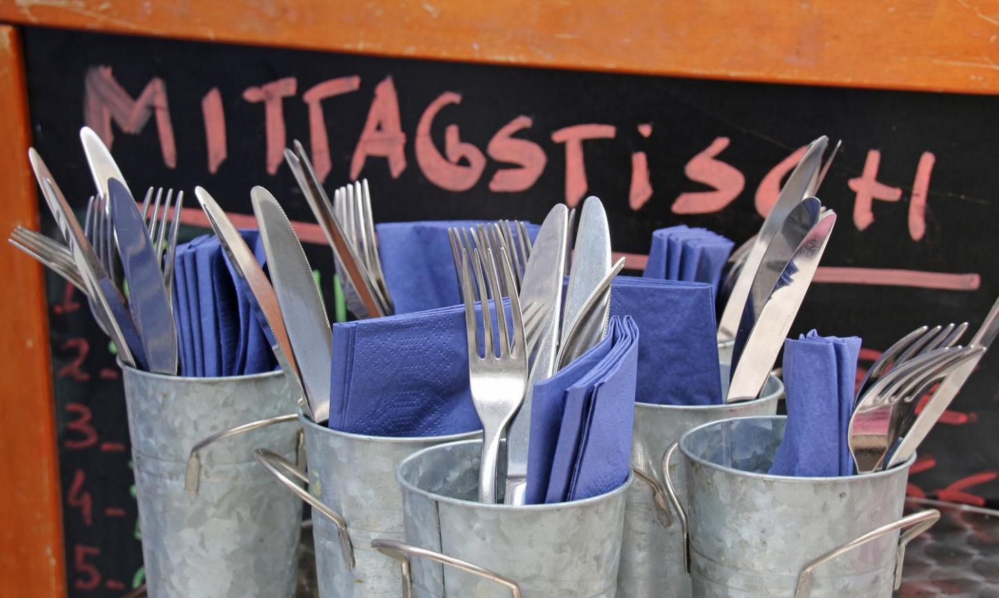 Mehrere Metallbecher sind mit Besteck und blauen Servietten gefüllt, davor steht eine Tafel mit der Aufschrift „Mittagstisch“.