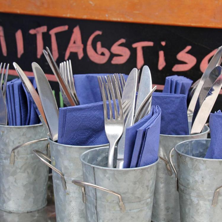 Mehrere Metallbecher sind mit Besteck und blauen Servietten gefüllt, davor steht eine Tafel mit der Aufschrift „Mittagstisch“.