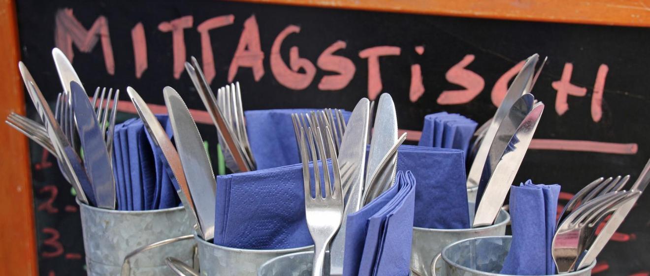 Mehrere Metallbecher sind mit Besteck und blauen Servietten gefüllt, davor steht eine Tafel mit der Aufschrift „Mittagstisch“.