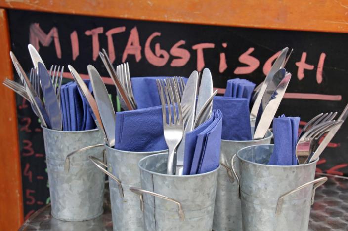 Mehrere Metallbecher sind mit Besteck und blauen Servietten gefüllt, davor steht eine Tafel mit der Aufschrift „Mittagstisch“.