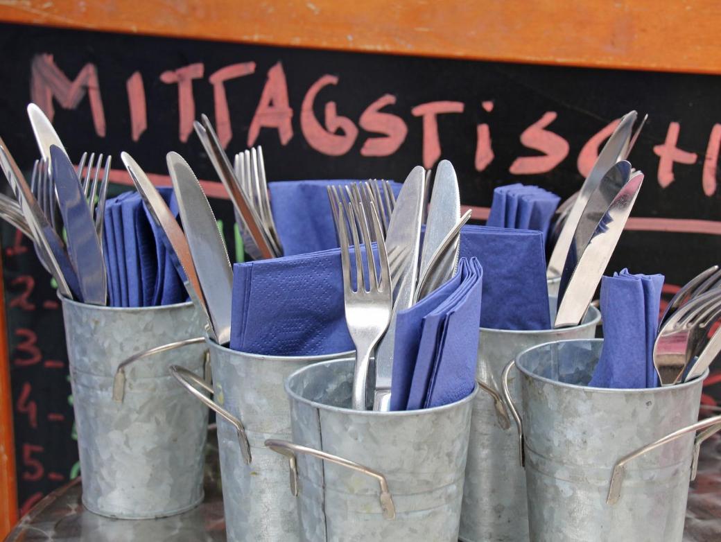 Mehrere Metallbecher sind mit Besteck und blauen Servietten gefüllt, davor steht eine Tafel mit der Aufschrift „Mittagstisch“.