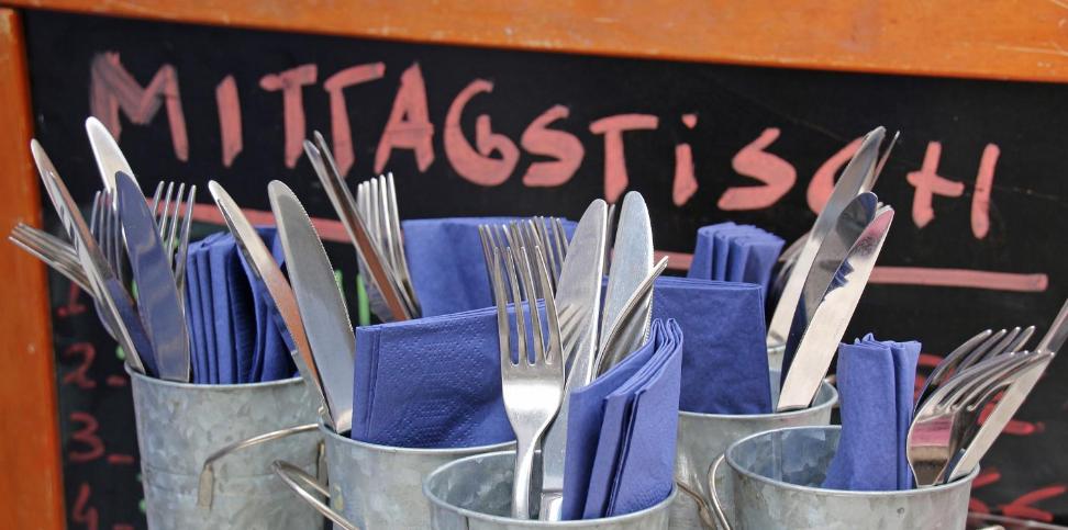 Mehrere Metallbecher sind mit Besteck und blauen Servietten gefüllt, davor steht eine Tafel mit der Aufschrift „Mittagstisch“.