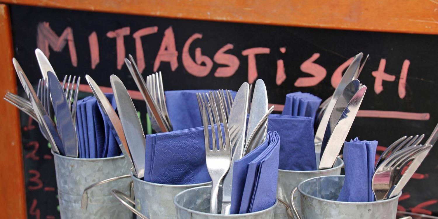 Mehrere Metallbecher sind mit Besteck und blauen Servietten gefüllt, davor steht eine Tafel mit der Aufschrift „Mittagstisch“.