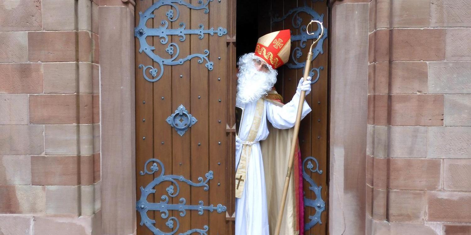 Das Bild zeigt eine Person in einem weißen Gewand und rotem Bischofshut, die mit einem Stab in der Hand durch eine halb geöffnete, reich verzierte Holztür einer Kirche tritt.