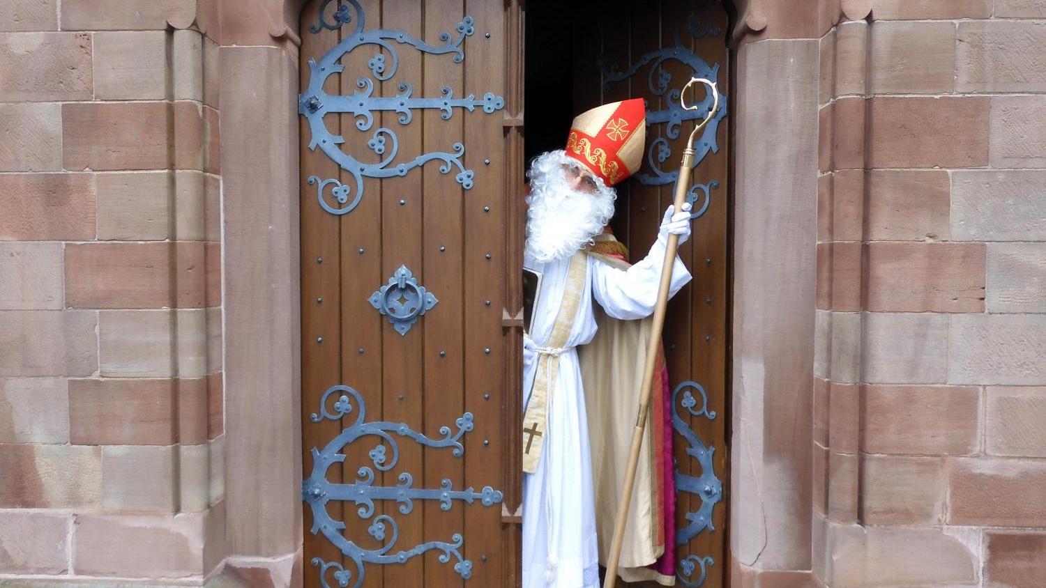 Das Bild zeigt eine Person in einem weißen Gewand und rotem Bischofshut, die mit einem Stab in der Hand durch eine halb geöffnete, reich verzierte Holztür einer Kirche tritt.