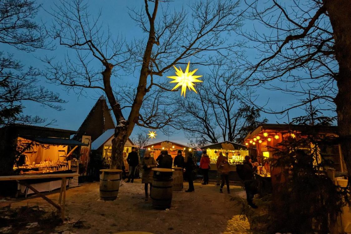 Das Foto zeigt einen stimmungsvollen Weihnachtsmarkt in der Abenddämmerung mit beleuchteten Holzständen, kahlen Bäumen und großen leuchtenden Sternen, die über den Besucherinnen und Besuchern hängen.