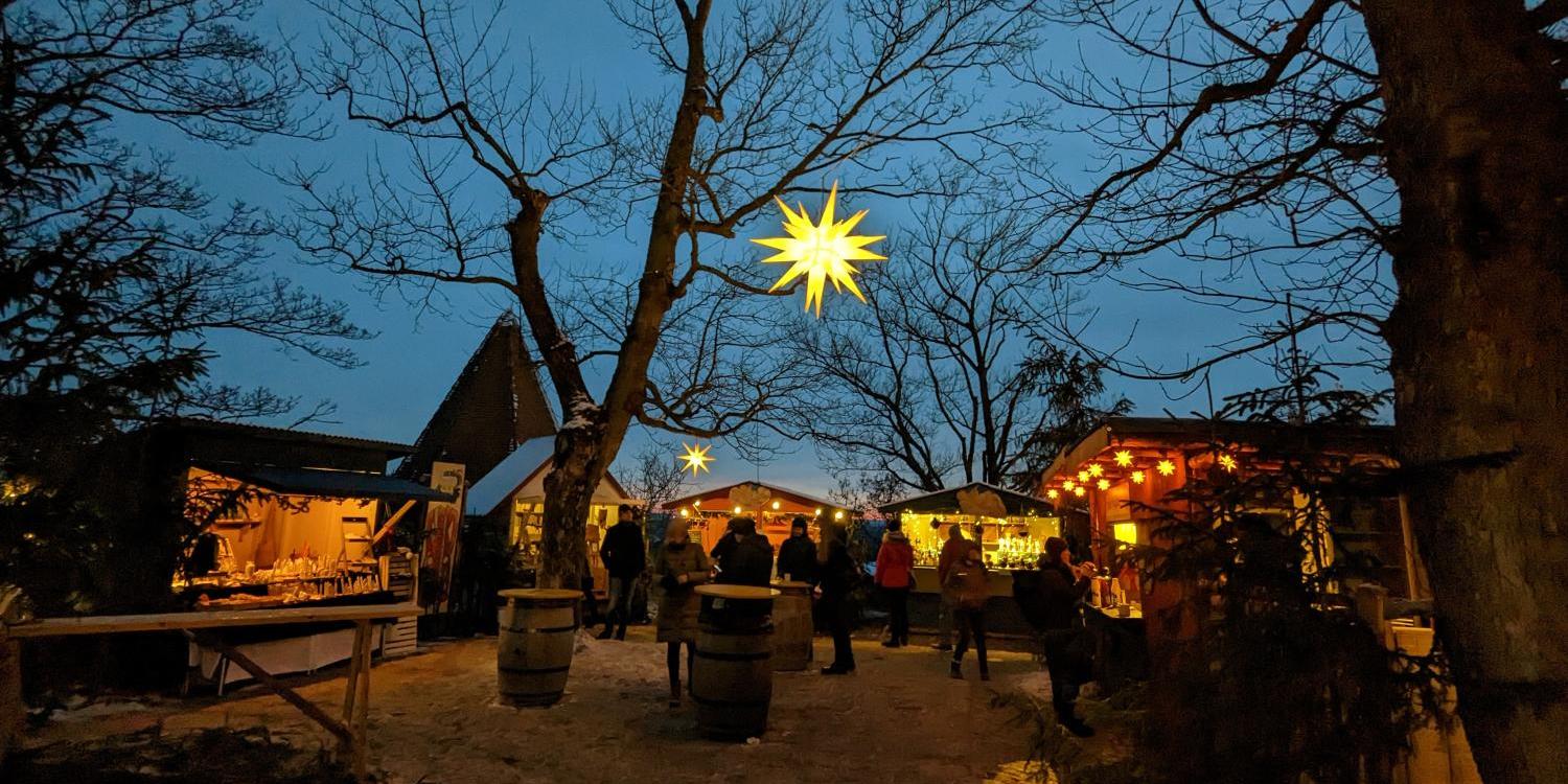 Das Foto zeigt einen stimmungsvollen Weihnachtsmarkt in der Abenddämmerung mit beleuchteten Holzständen, kahlen Bäumen und großen leuchtenden Sternen, die über den Besucherinnen und Besuchern hängen.