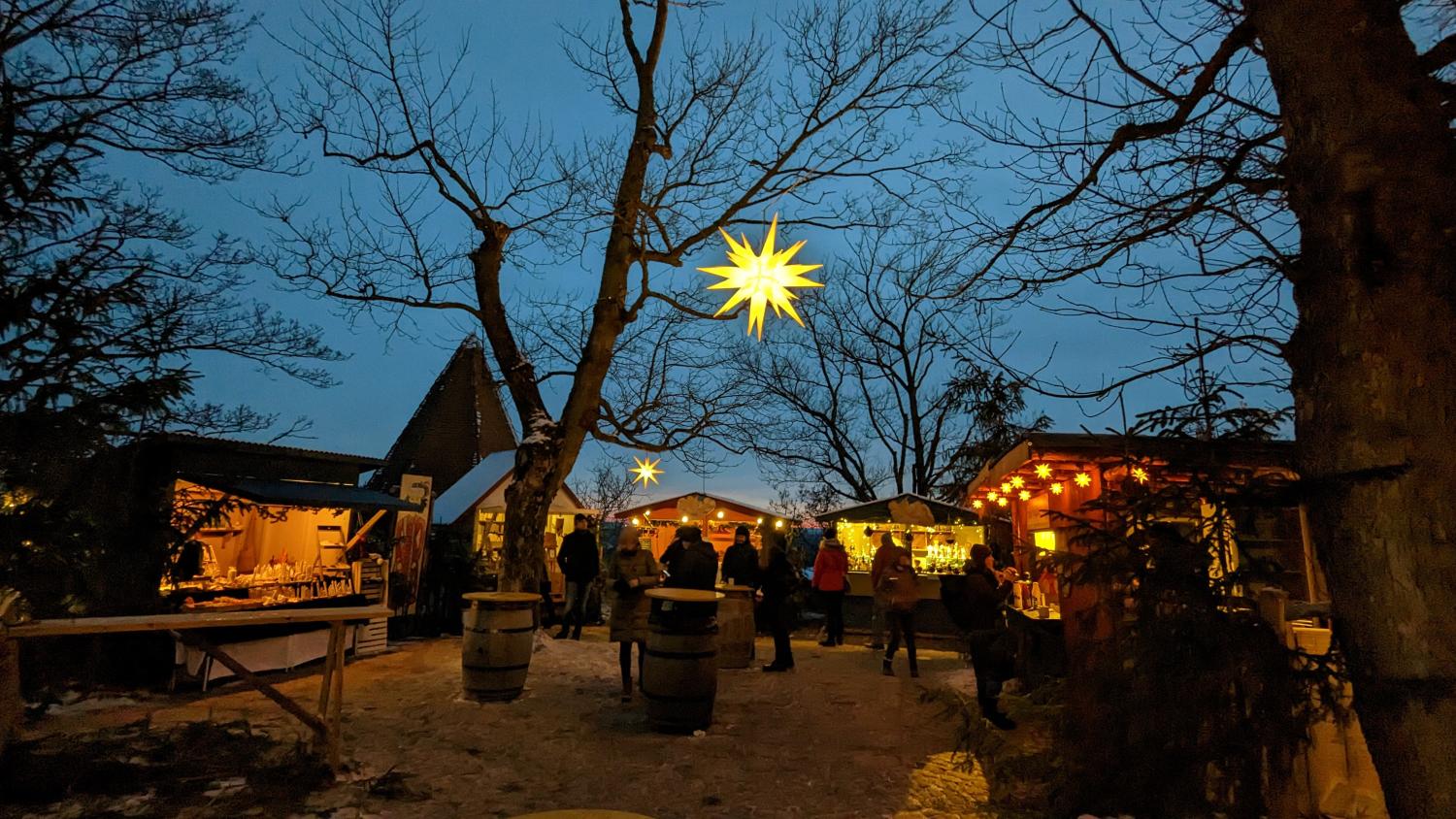 Das Foto zeigt einen stimmungsvollen Weihnachtsmarkt in der Abenddämmerung mit beleuchteten Holzständen, kahlen Bäumen und großen leuchtenden Sternen, die über den Besucherinnen und Besuchern hängen.