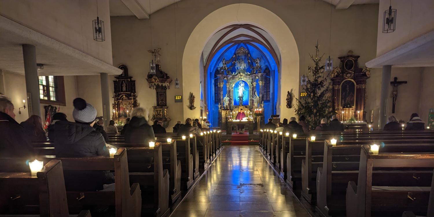 Das Foto zeigt eine festlich beleuchtete Kirche im Advent, in der Gläubige bei Kerzenschein einer Messe vor dem blau erleuchteten Altar beiwohnen.