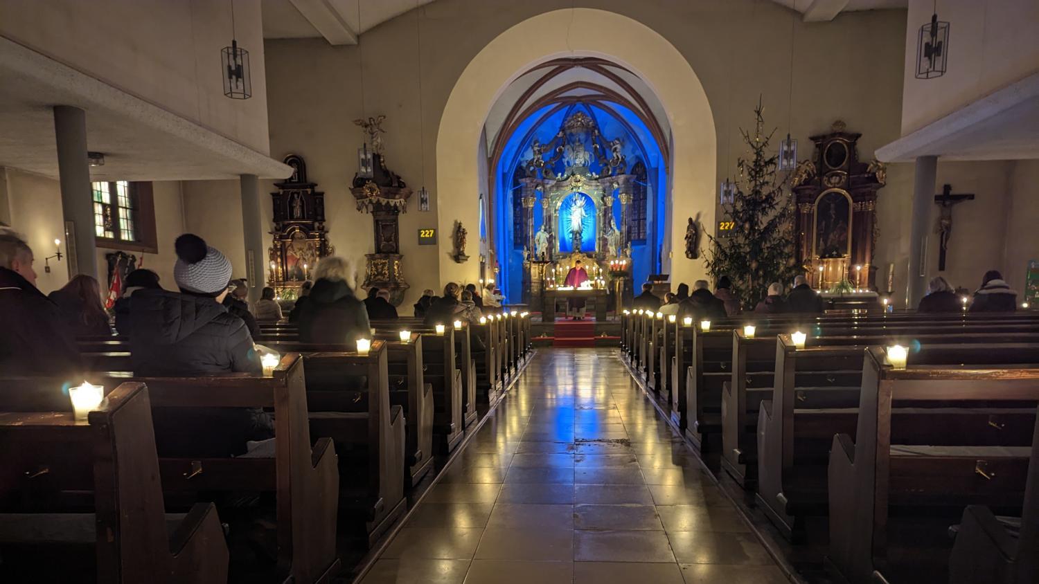 Das Foto zeigt eine festlich beleuchtete Kirche im Advent, in der Gläubige bei Kerzenschein einer Messe vor dem blau erleuchteten Altar beiwohnen.