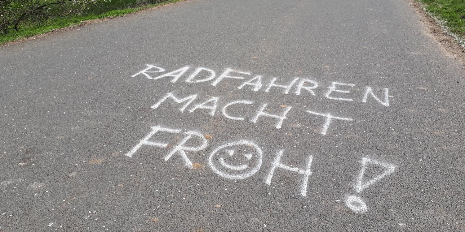 Text „Radfahren macht froh!“ auf asphaltiertem Radweg, blühende Bäume am Rand.