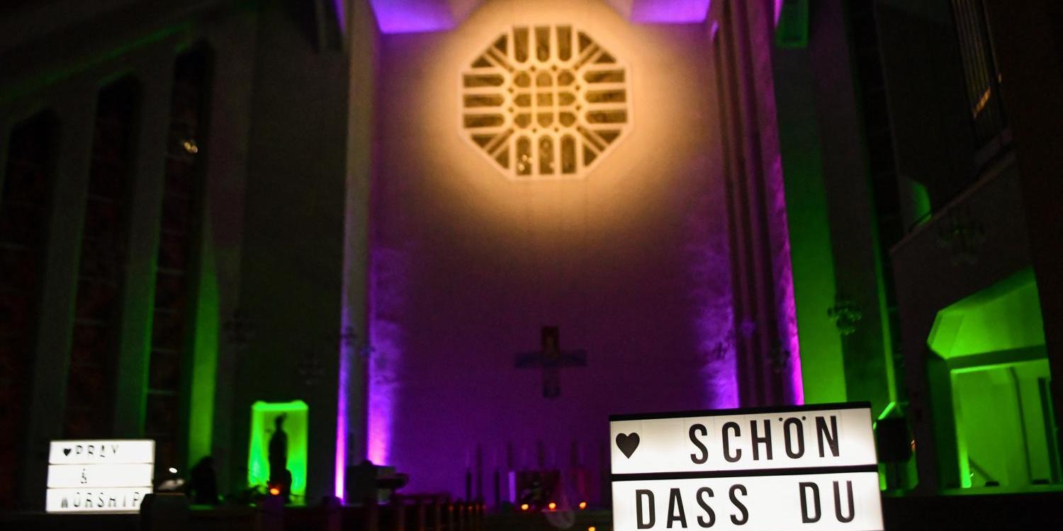 Das Foto zeigt den Innenraum einer Kirche, bunt in Grün und Violett beleuchtet, mit einem großen Lichtkasten im Vordergrund, auf dem die Worte „Schön dass du da bist“ stehen.