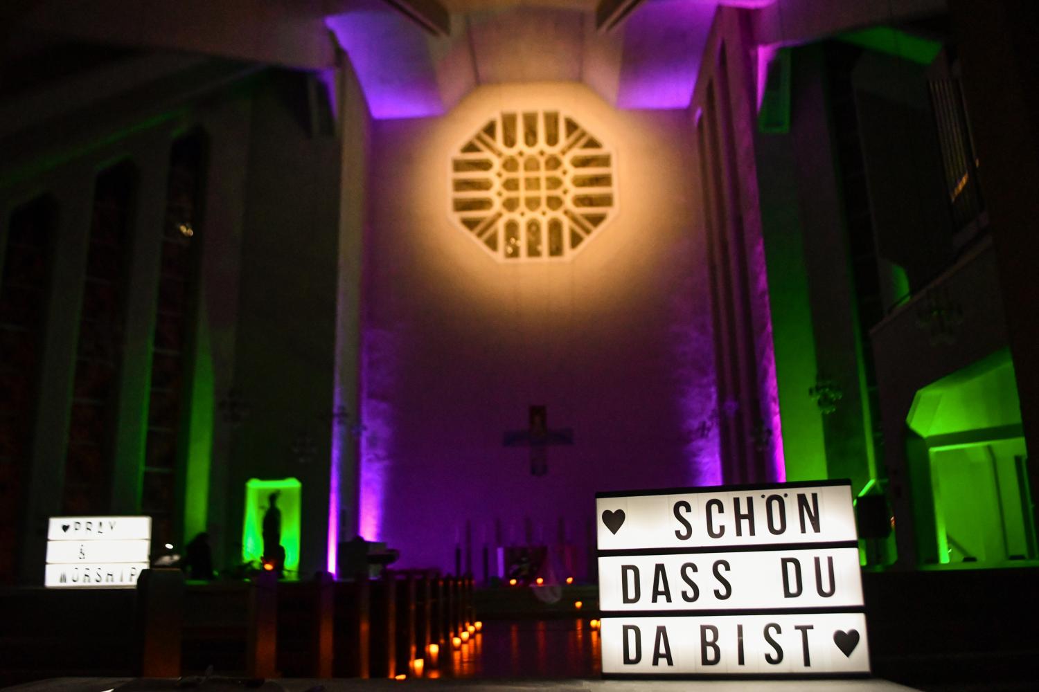 Das Foto zeigt den Innenraum einer Kirche, bunt in Grün und Violett beleuchtet, mit einem großen Lichtkasten im Vordergrund, auf dem die Worte „Schön dass du da bist“ stehen.