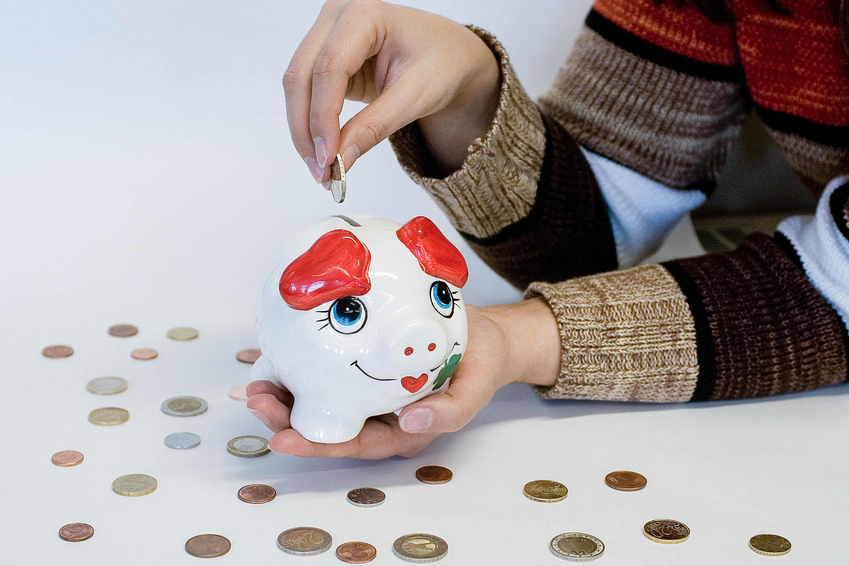 Eine Person steckt eine Münze in ein weißes Sparschwein mit roten Ohren und blauen Augen, umgeben von verstreuten Münzen auf weißem Hintergrund.