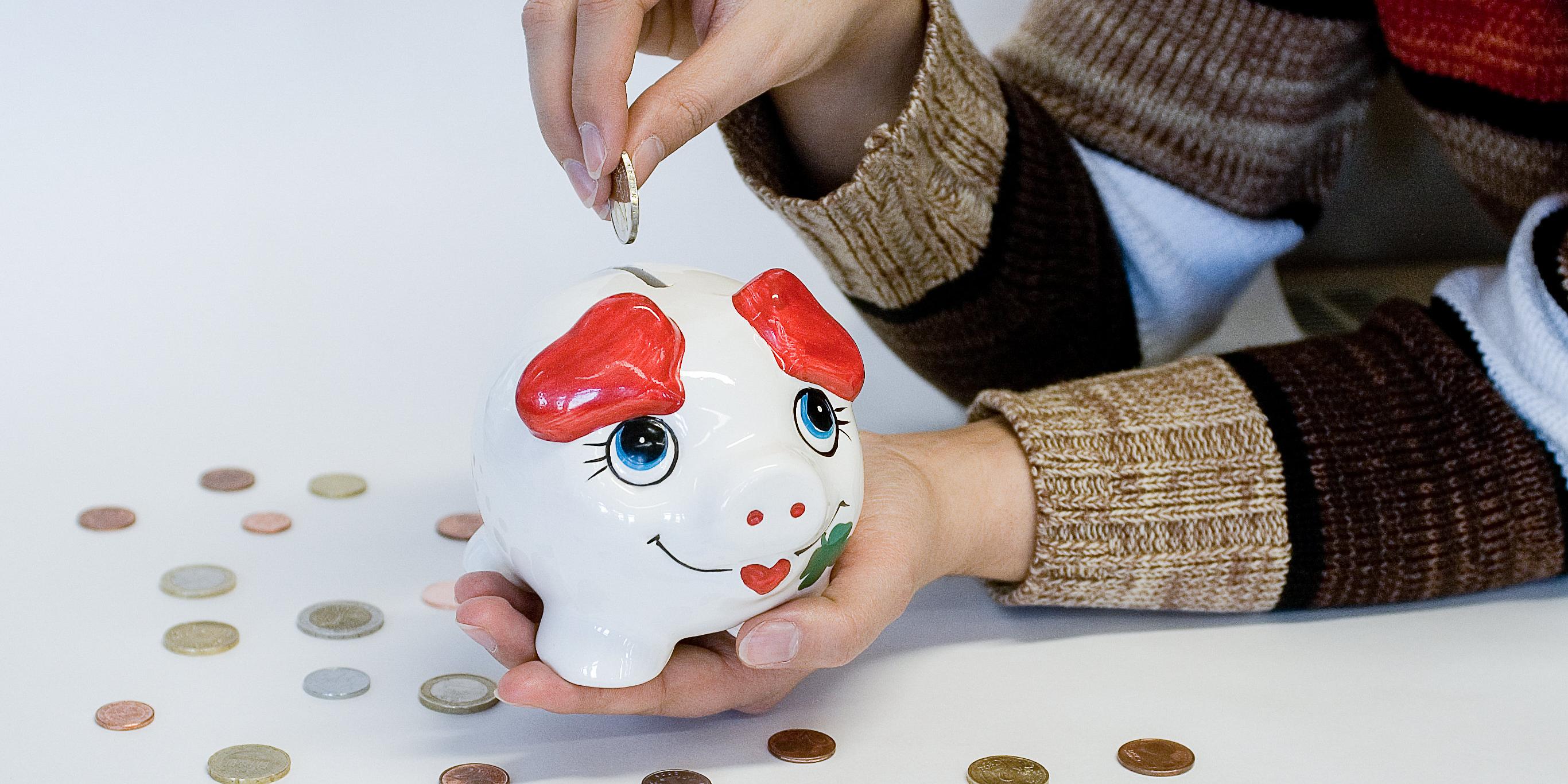 Eine Person steckt eine Münze in ein weißes Sparschwein mit roten Ohren und blauen Augen, umgeben von verstreuten Münzen auf weißem Hintergrund.
