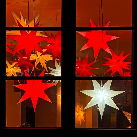 Das Bild zeigt ein Fenster, vor dem mehrere leuchtende Sterne in Rot, Gelb und Weiß an Schnüren hängen und eine warme, festliche Stimmung erzeugen.