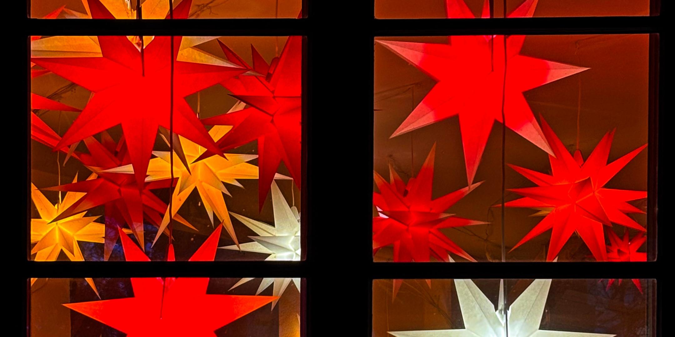 Das Bild zeigt ein Fenster, vor dem mehrere leuchtende Sterne in Rot, Gelb und Weiß an Schnüren hängen und eine warme, festliche Stimmung erzeugen.