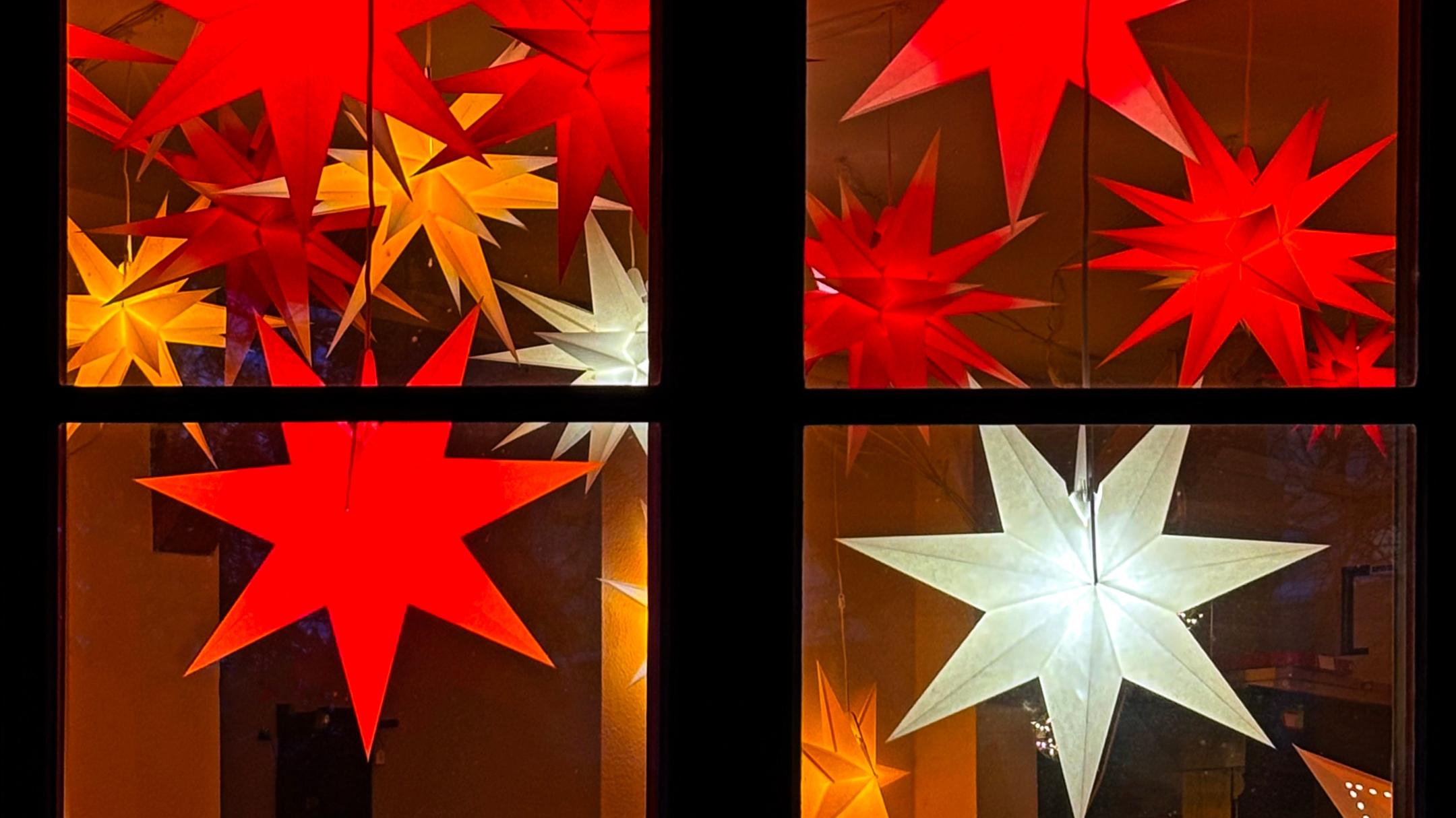 Das Bild zeigt ein Fenster, vor dem mehrere leuchtende Sterne in Rot, Gelb und Weiß an Schnüren hängen und eine warme, festliche Stimmung erzeugen.