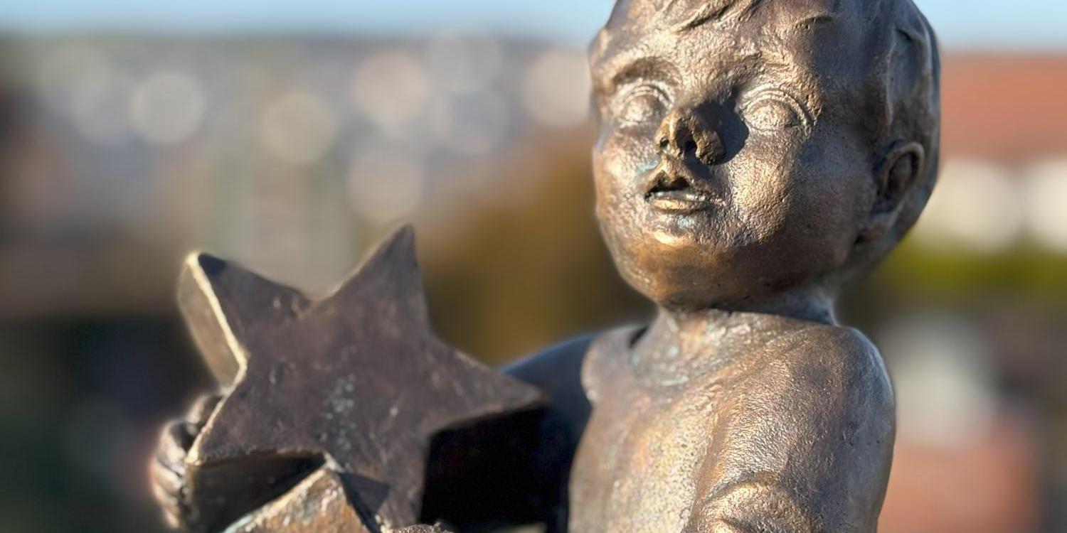 Eine bronzefarbene Skulptur eines sitzenden Kindes, das mehrere sternförmige Figuren in den Händen hält.