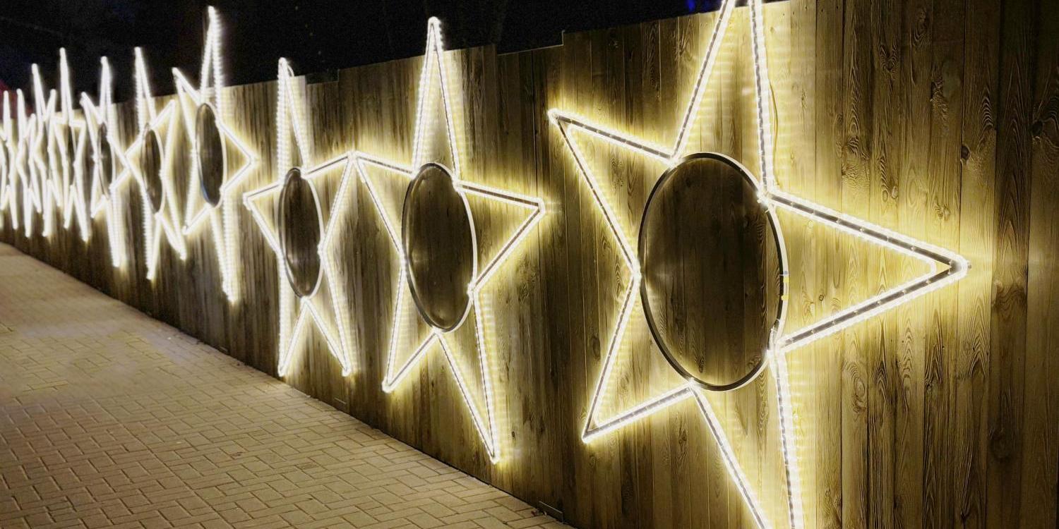 Eine Reihe großer, leuchtender Sterne aus weißen Lichtschläuchen ist an einem hölzernen Zaun befestigt und erhellt den gepflasterten Weg in der Dunkelheit.