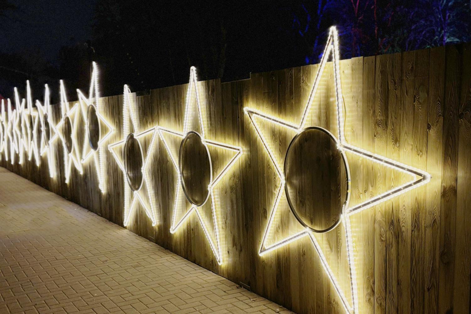 Eine Reihe großer, leuchtender Sterne aus weißen Lichtschläuchen ist an einem hölzernen Zaun befestigt und erhellt den gepflasterten Weg in der Dunkelheit.