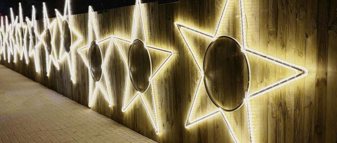 Eine Reihe großer, leuchtender Sterne aus weißen Lichtschläuchen ist an einem hölzernen Zaun befestigt und erhellt den gepflasterten Weg in der Dunkelheit.