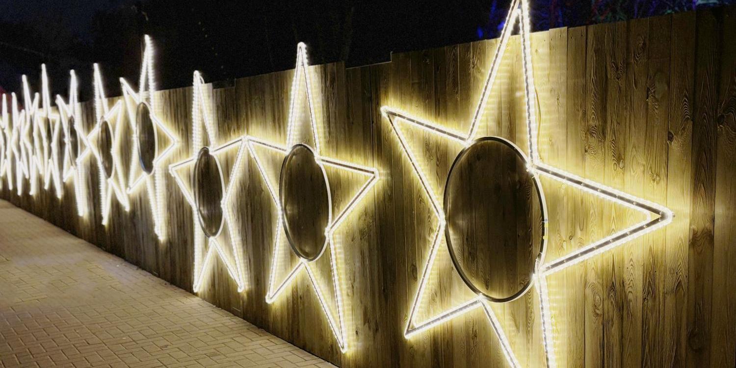 Eine Reihe großer, leuchtender Sterne aus weißen Lichtschläuchen ist an einem hölzernen Zaun befestigt und erhellt den gepflasterten Weg in der Dunkelheit.