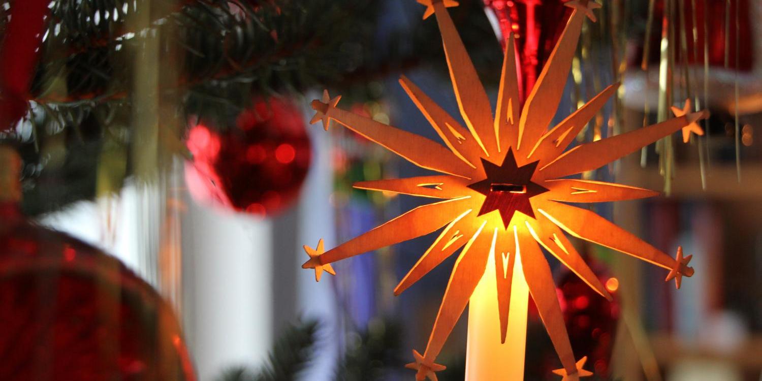 Ein leuchtender, goldener Papierstern strahlt warmes Licht zwischen roten Weihnachtskugeln und Tannenzweigen.