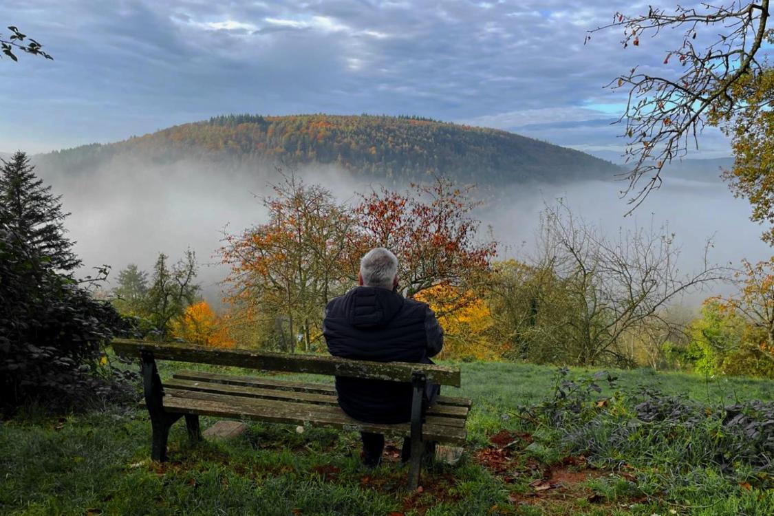 Ein Mensch sitzt auf einer Bank und blickt still über eine nebelverhangene, herbstlich gefärbte Landschaft mit Hügeln und farbigen Bäumen.