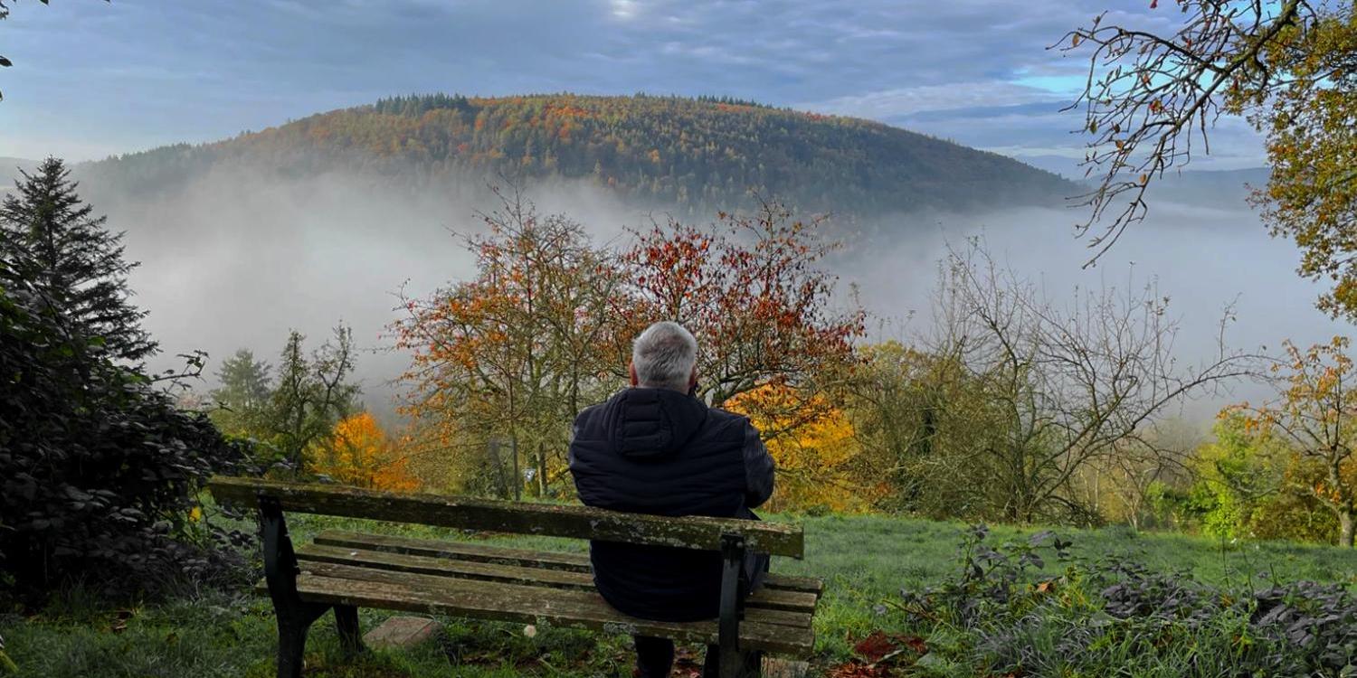 Ein Mensch sitzt auf einer Bank und blickt still über eine nebelverhangene, herbstlich gefärbte Landschaft mit Hügeln und farbigen Bäumen.