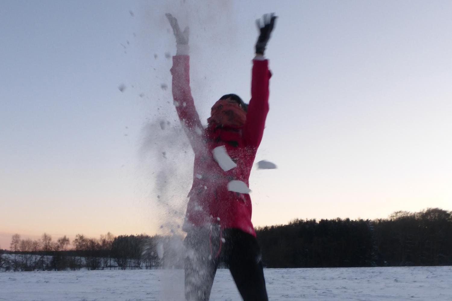 Eine Person steht in einer verschneiten Landschaft und wirft Schnee mit erhobenen Armen in die Luft, sodass eine weiße Wolke aus Schnee um sie herum aufsteigt.