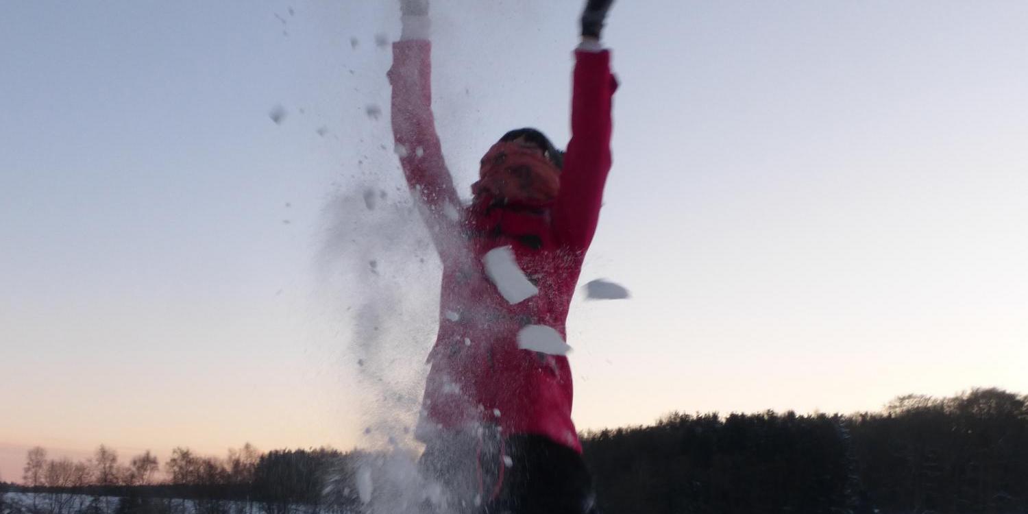Eine Person steht in einer verschneiten Landschaft und wirft Schnee mit erhobenen Armen in die Luft, sodass eine weiße Wolke aus Schnee um sie herum aufsteigt.