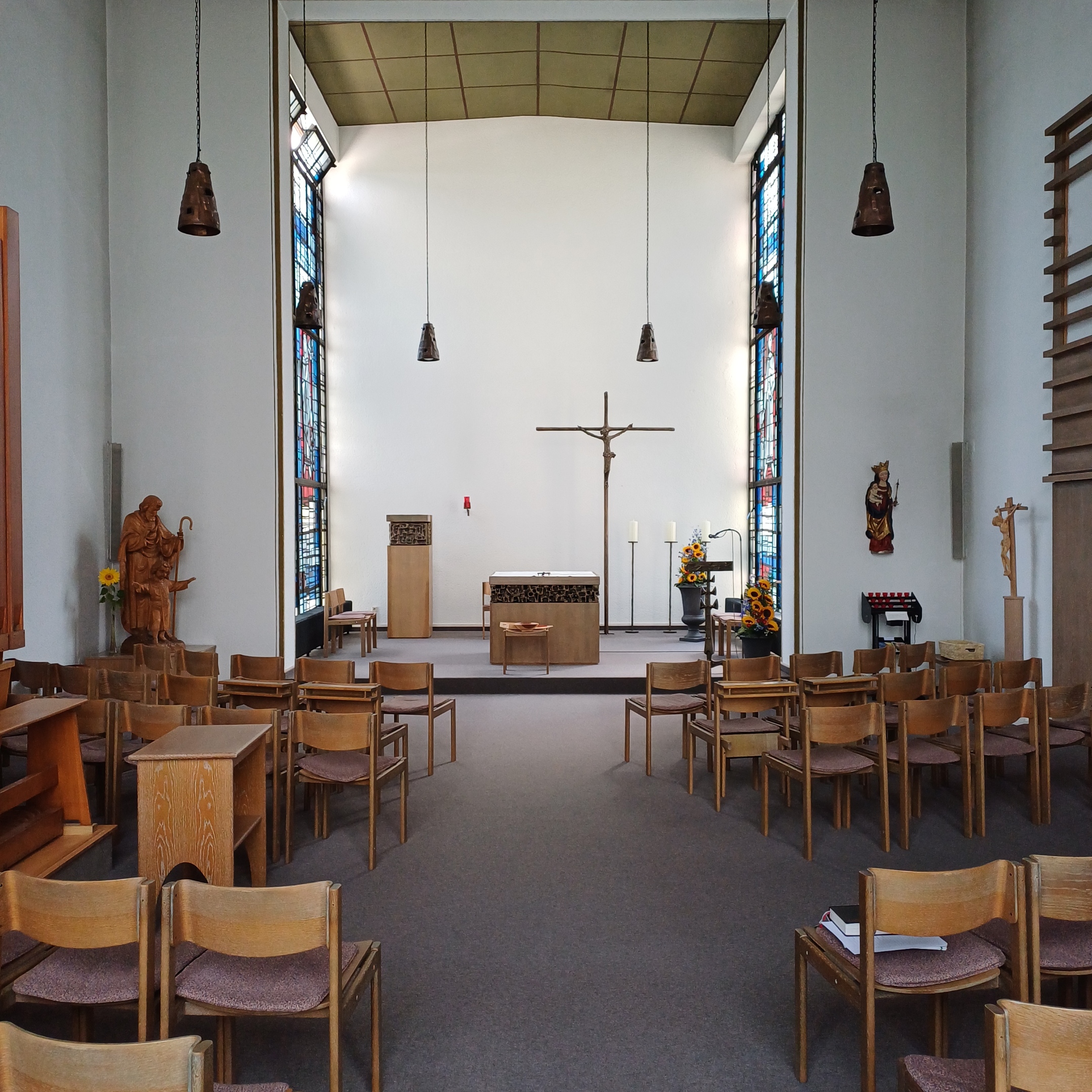 Das Bild zeigt das Innere einer Kapelle mit einem Mittelgang, Holzstühlen, einem Altar mit Kruzifix, Sonnenblumen, Kerzenständern und bunten Glasfenstern, flankiert von religiösen Statuen und dekorativen Elementen.