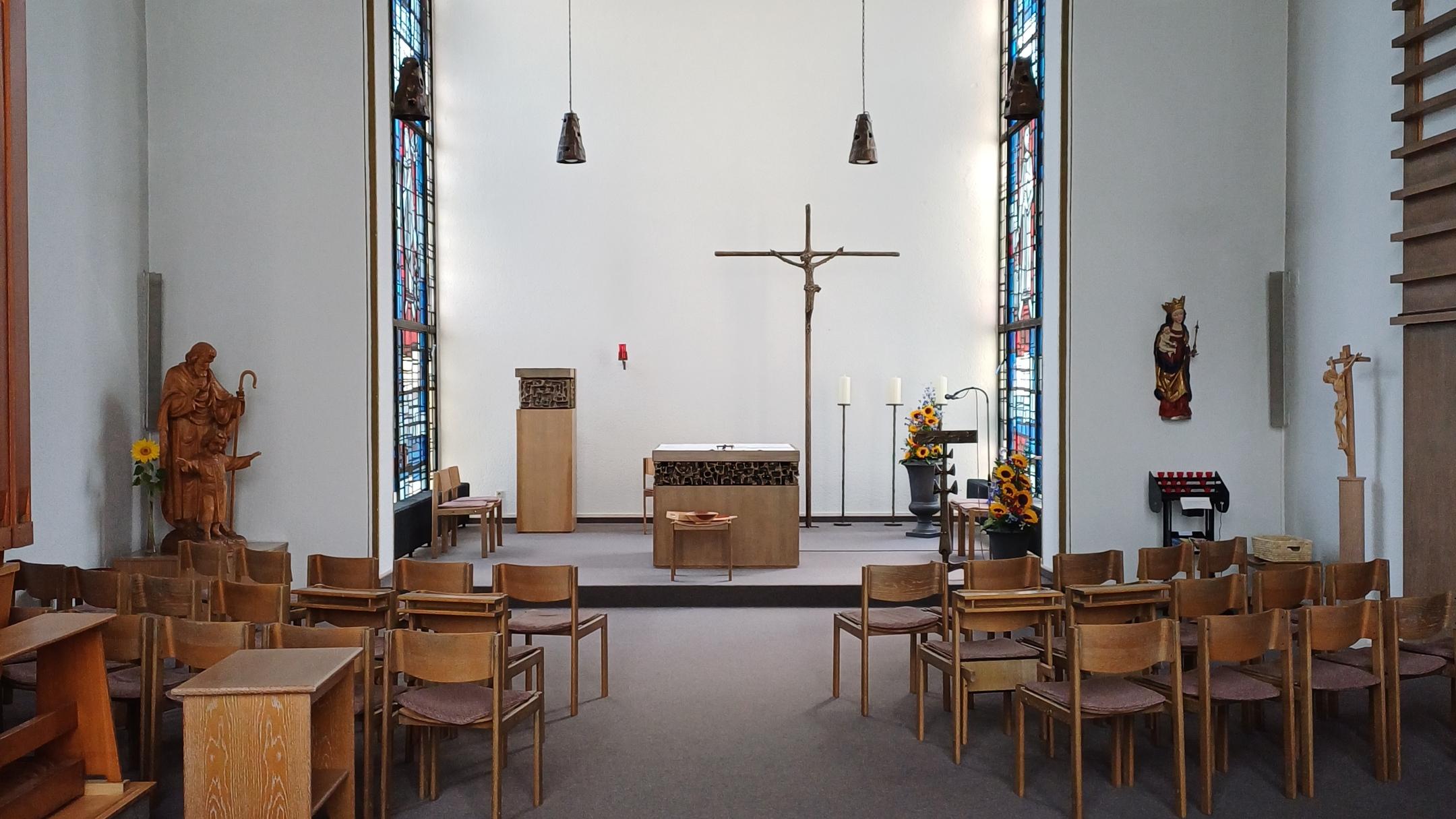 Das Bild zeigt das Innere einer Kapelle mit einem Mittelgang, Holzstühlen, einem Altar mit Kruzifix, Sonnenblumen, Kerzenständern und bunten Glasfenstern, flankiert von religiösen Statuen und dekorativen Elementen.
