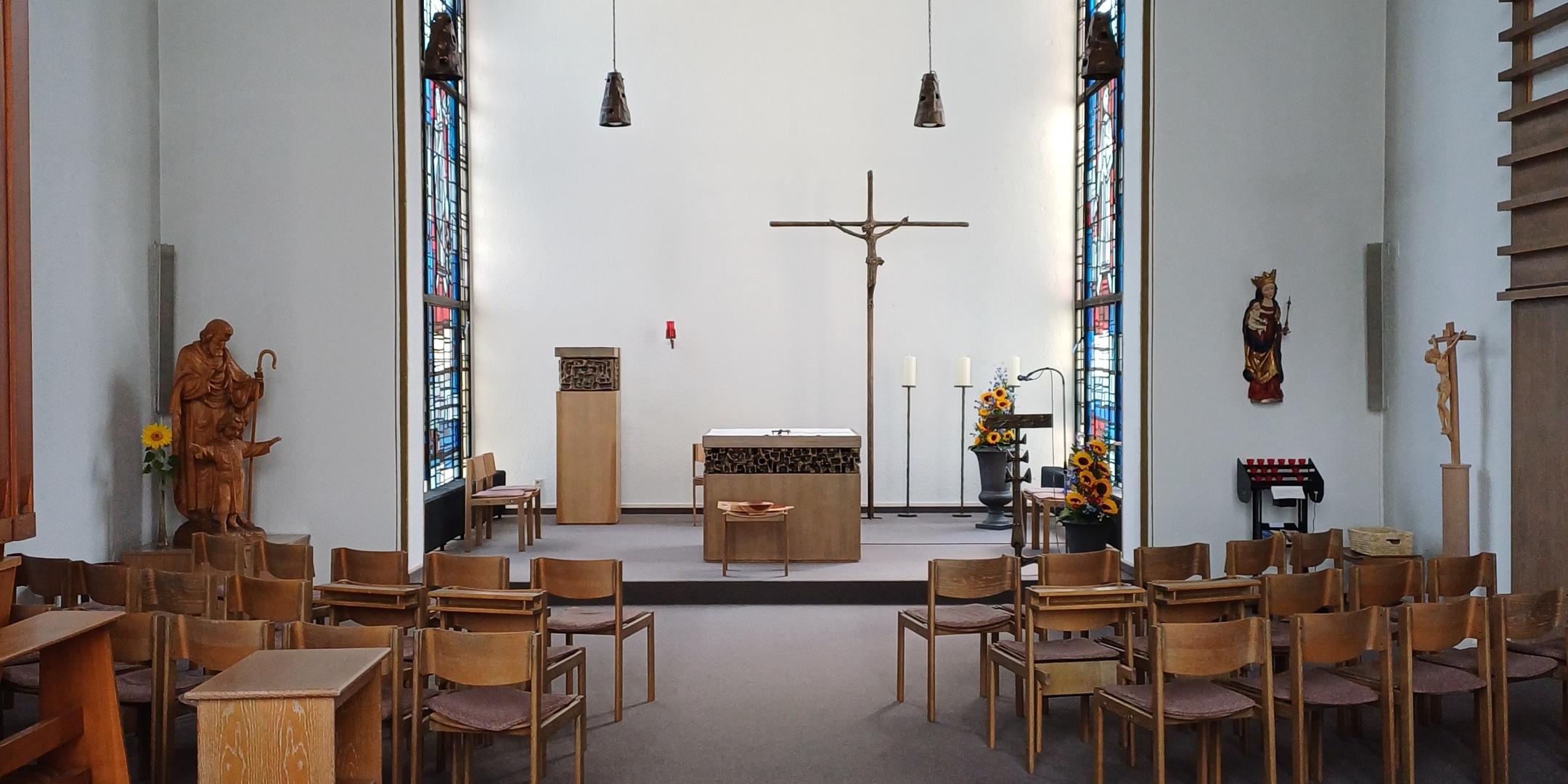 Das Bild zeigt das Innere einer Kapelle mit einem Mittelgang, Holzstühlen, einem Altar mit Kruzifix, Sonnenblumen, Kerzenständern und bunten Glasfenstern, flankiert von religiösen Statuen und dekorativen Elementen.
