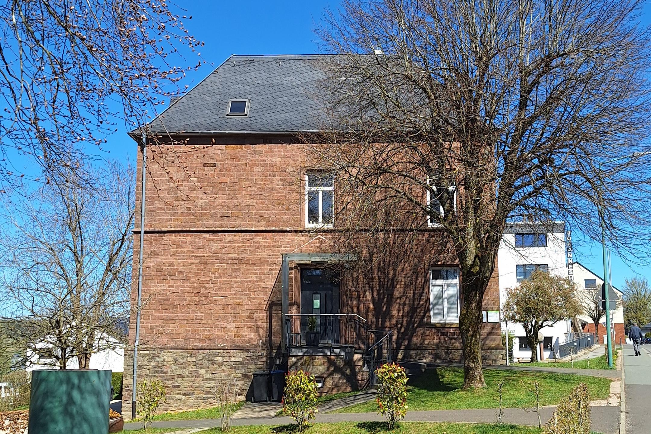 Ein Backsteingebäude an einer ruhigen Dorfstraße, umgeben von Bäumen und Grünflächen