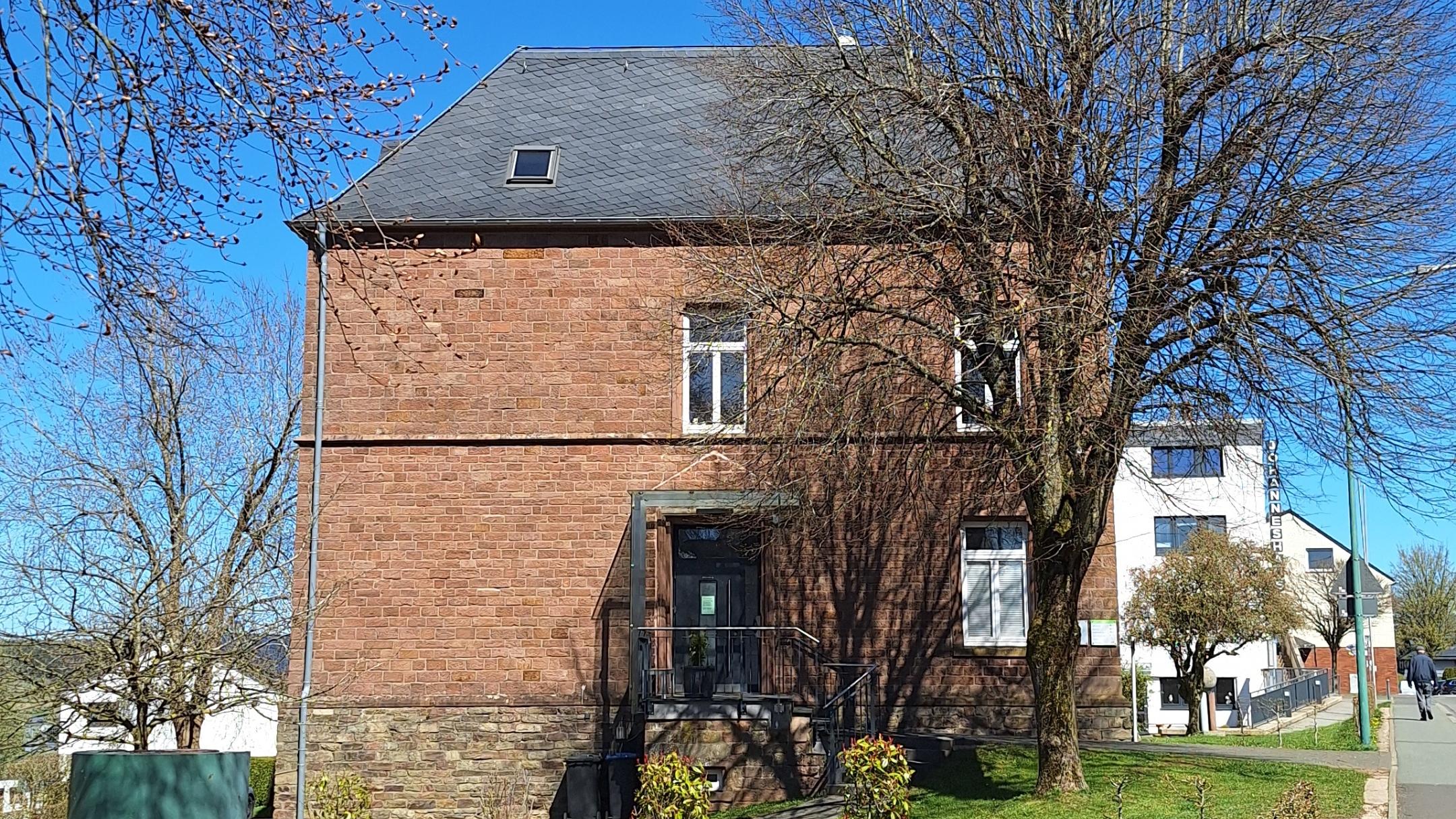 Ein Backsteingebäude an einer ruhigen Dorfstraße, umgeben von Bäumen und Grünflächen