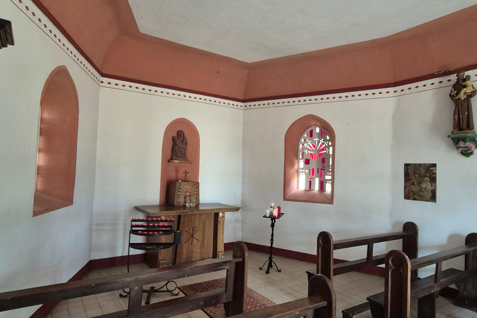 Der Innenraum der St.-Anna-Kapelle mit Holzbänken, einem schlichten Altar, einer Pietà in einer Wandnische und einem farbigen Fenster.
