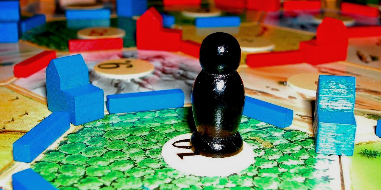 Das Foto zeigt ein Spielbrett mit sechseckigen Feldern, bunten Holzspielsteinen und einer schwarzen Spielfigur auf einem grünen Feld mit der Zahl 10.