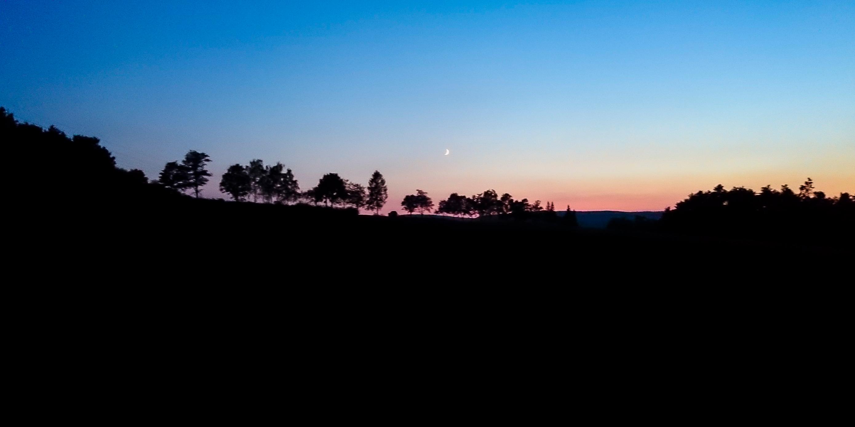 Das Bild zeigt eine ruhige Landschaft in der Dämmerung mit einem farbenprächtigen Himmel, der von tiefem Blau zu warmem Orange übergeht. Vor dem Horizont zeichnen sich dunkle Baum-Silhouetten ab.