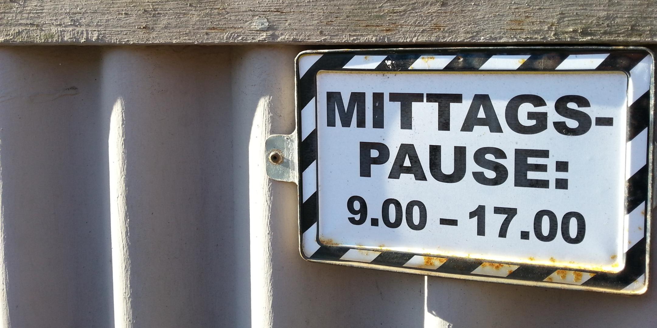 Das Foto zeigt ein Schild mit der Aufschrift „MITTAGSPAUSE: 9.00 – 17.00“, befestigt an einem Metallgeländer vor einem Gebäude mit Holz- und Steinfassade.
