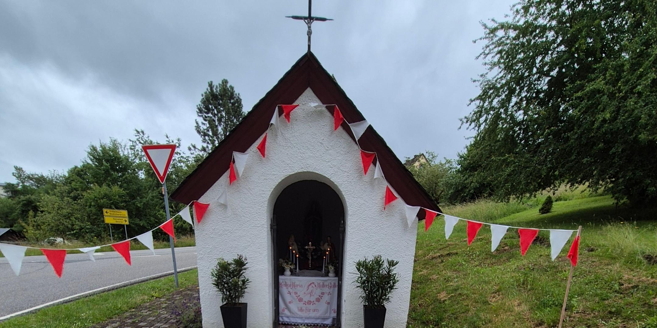 Kleine weiße Kapelle mit Kreuz auf dem Dach, geschmückt mit roten und weißen Wimpeln im Grünen.